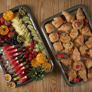 Two trays of food on a wooden surface, one with fruits and the other with pastries.