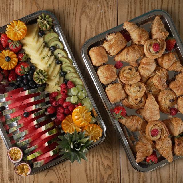 Two trays of food on a wooden surface, one with fruits and the other with pastries.